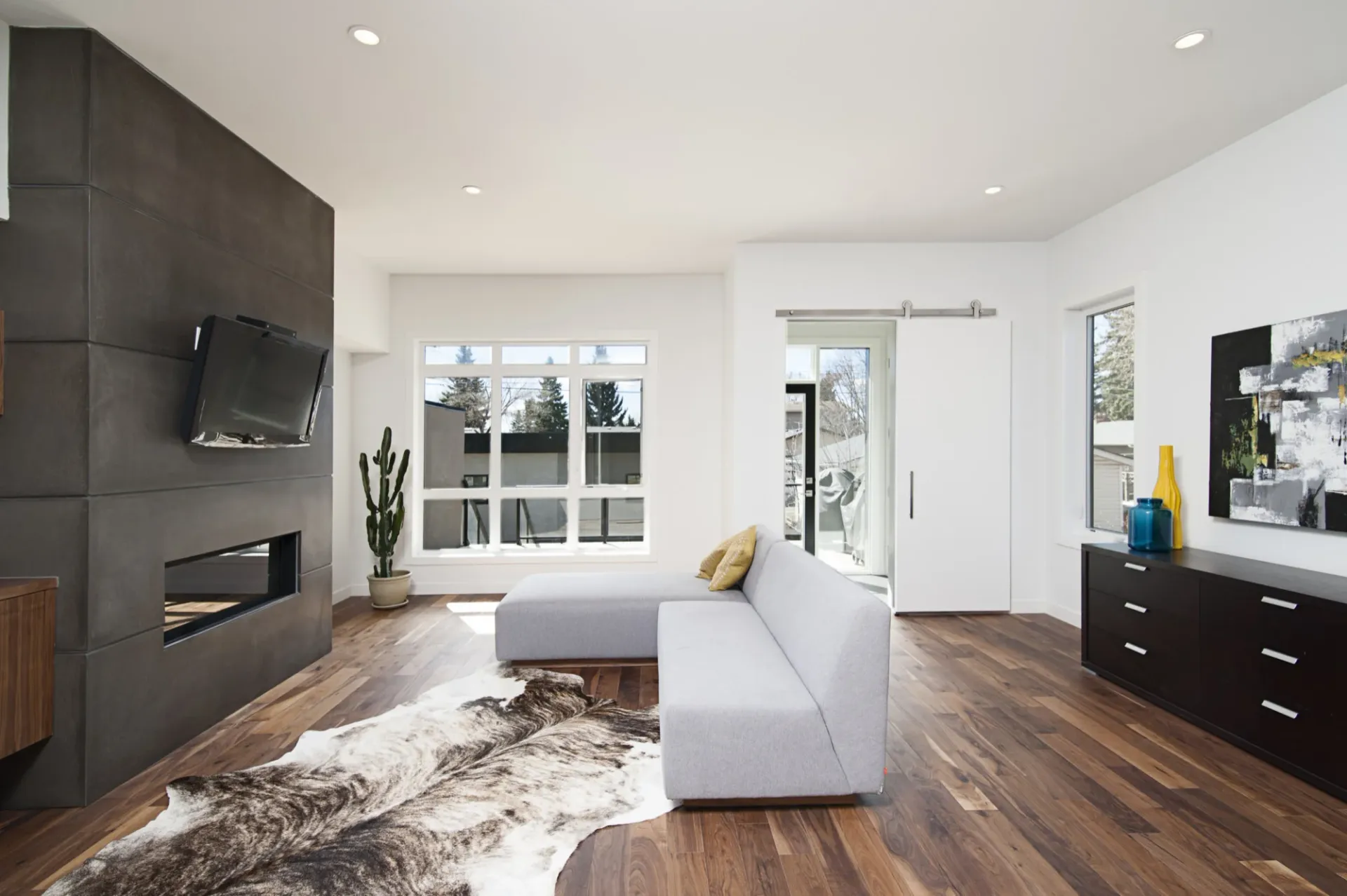A photo of a living room with wood floors, a cowhide rug, a dark wooden accent wall with a fireplace and a TV mounted to the wall on the left. A modern light gray sectional couch is towards the right.