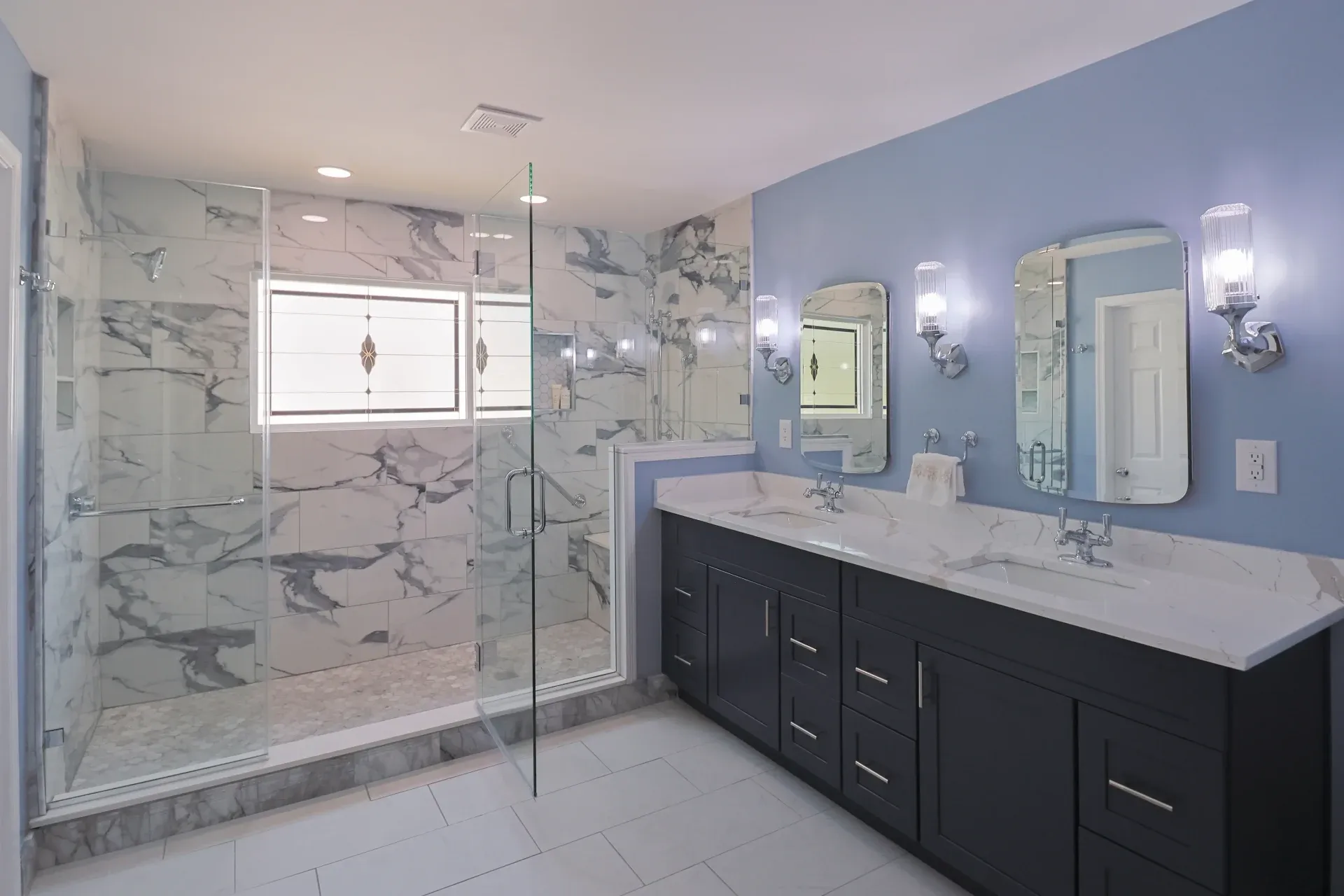 A photo of a bathroom with a large, glass-doored marble shower with a matching double vanity with dark cabinets.